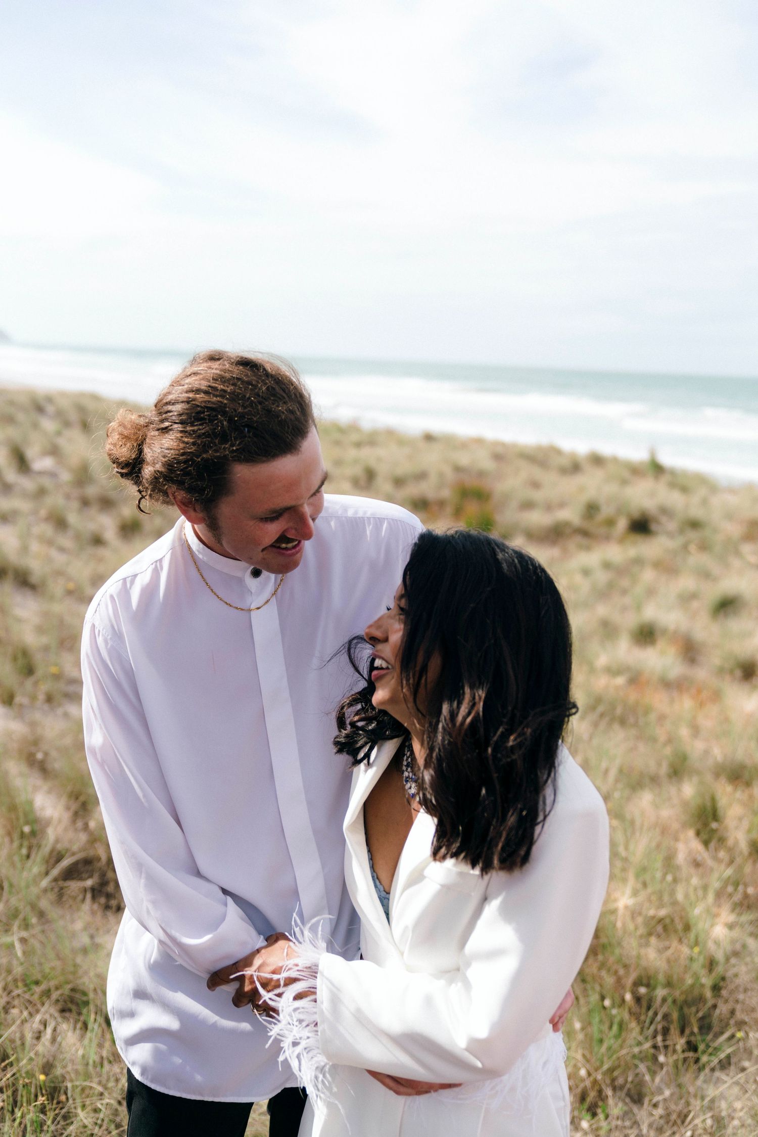 Romantic couple in white embracing on windswept coastal dunes with ocean backdrop.