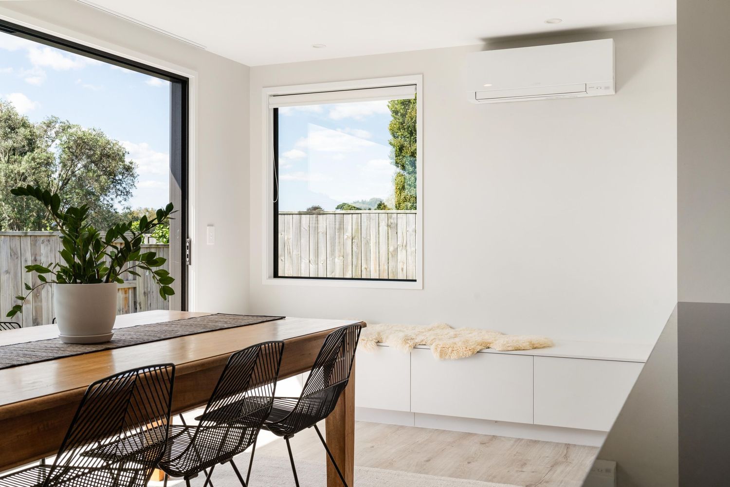 Modern minimalist dining area with long wooden table, black chairs and bright white walls with natural light streaming in.
