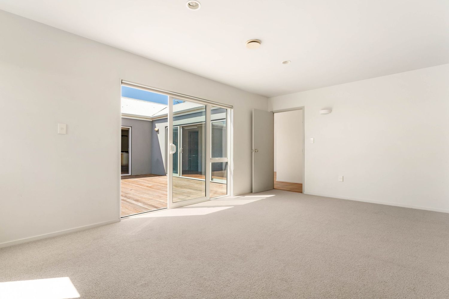 Bright empty room with carpet flooring and glass sliding doors opening to an outdoor courtyard.
