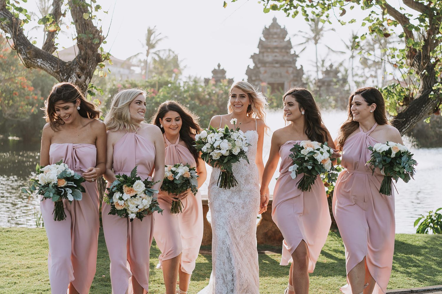 Bridesmaids in blush pink dresses pose with bride holding bouquets in an outdoor garden setting.