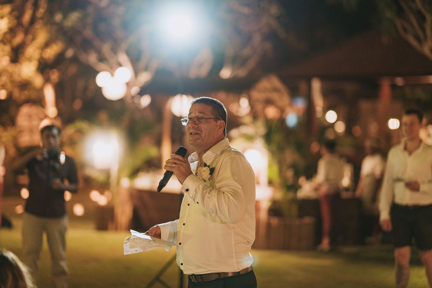 A nighttime outdoor celebration with bright spotlights illuminating trees and party tables.