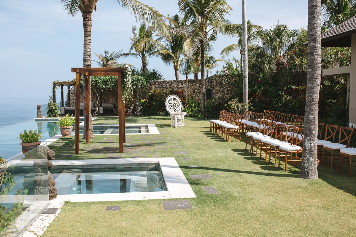 Outdoor wedding ceremony setup with wooden benches on grass lawn overlooking ocean, featuring pool and tropical palm trees.