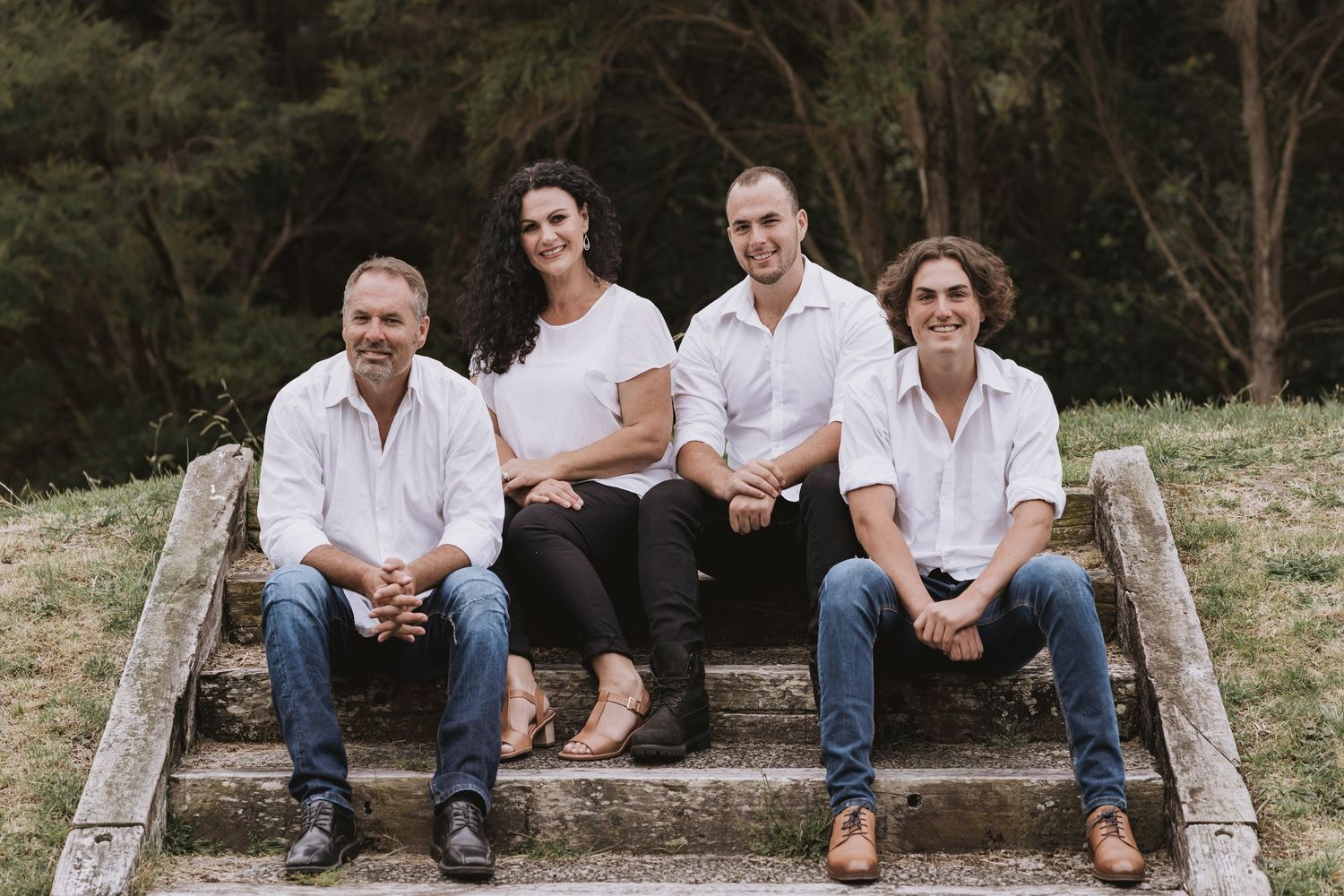 Four family members sit casually together on outdoor stone steps against a natural wooded backdrop.