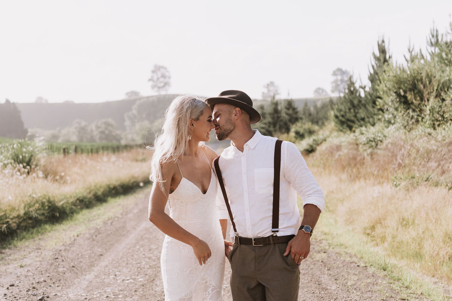 Newlywed couple walking together on a rustic dirt road with mountains and fields in the background.