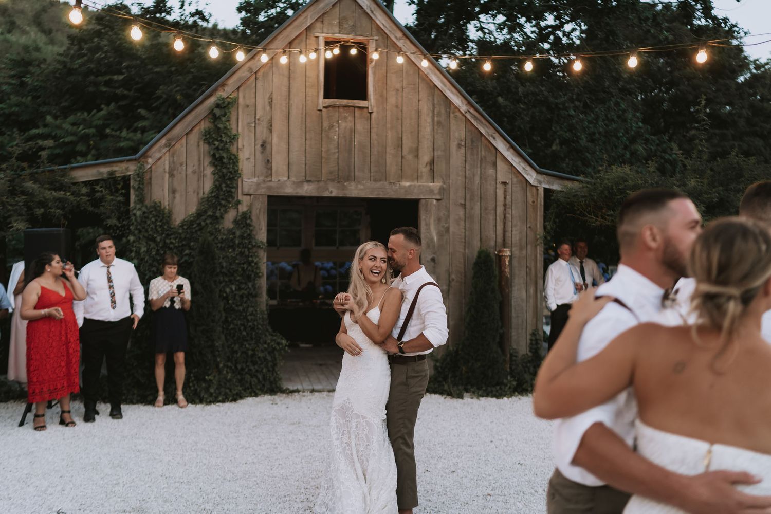 A rustic barn wedding celebration with people dancing and embracing on a white ground at sunset.