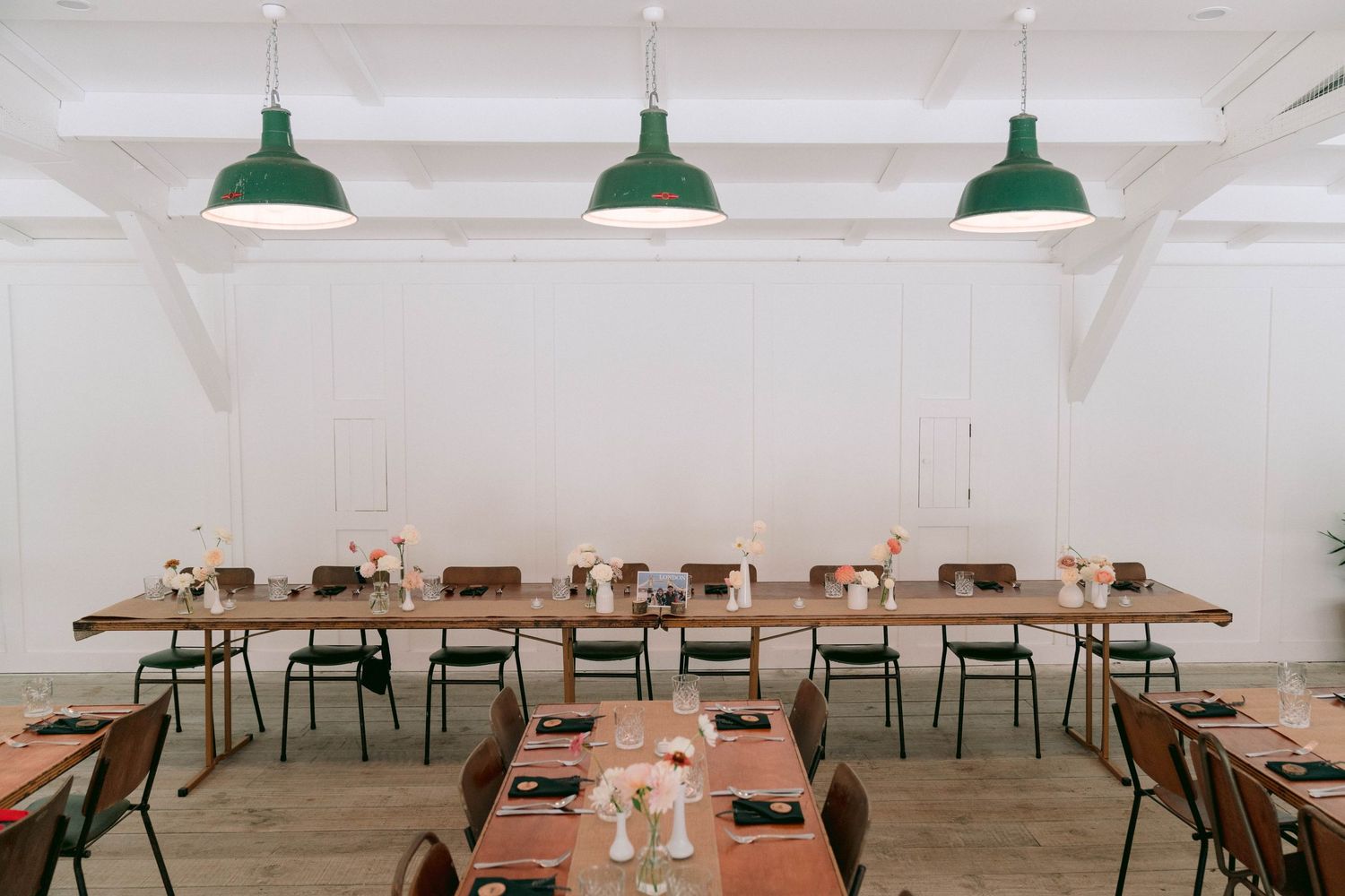 Modern dining room with green pendant lights, long wooden tables, and metal chairs in a bright white space.