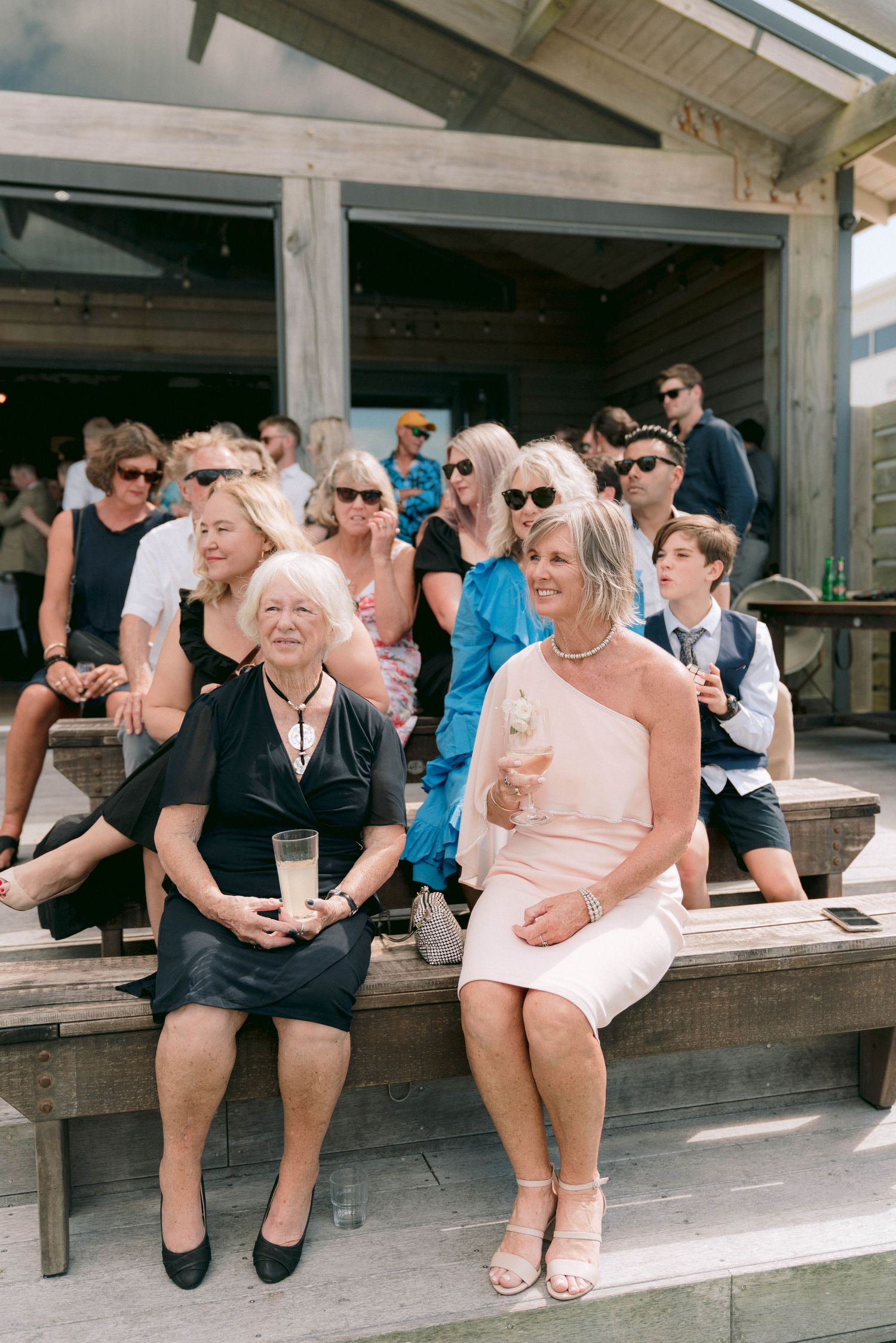 Wedding guests sit and laugh together on a rustic wooden bench during an outdoor celebration.