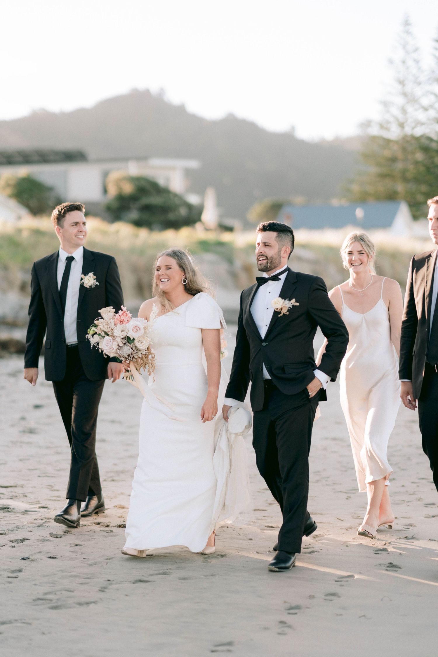 Wedding party walks along sandy beach in formal attire during sunset photoshoot.