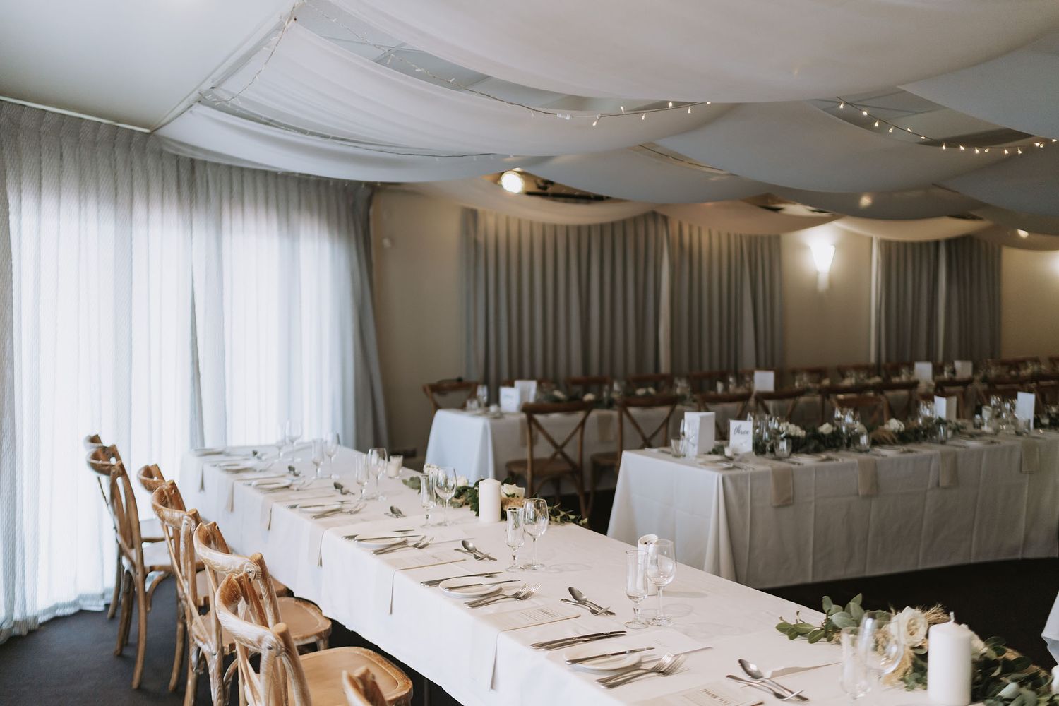 Elegant wedding reception venue with long wooden tables and white linens set for dining under soft overhead lighting.