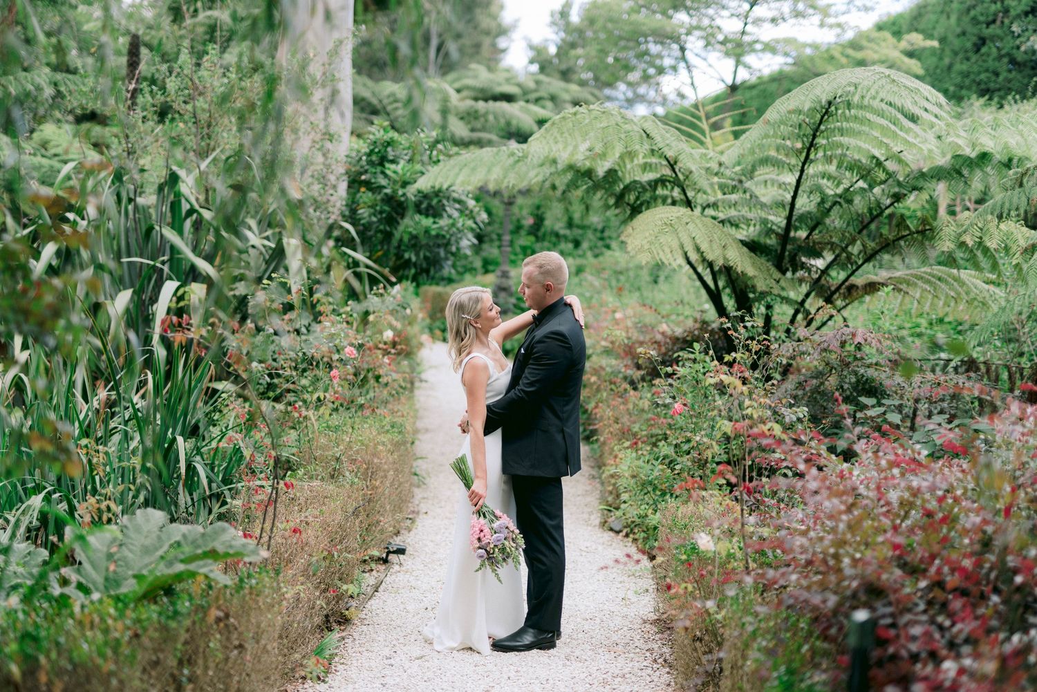 A couple celebrates their wedding surrounded by lush garden foliage and flowering plants along a winding path.