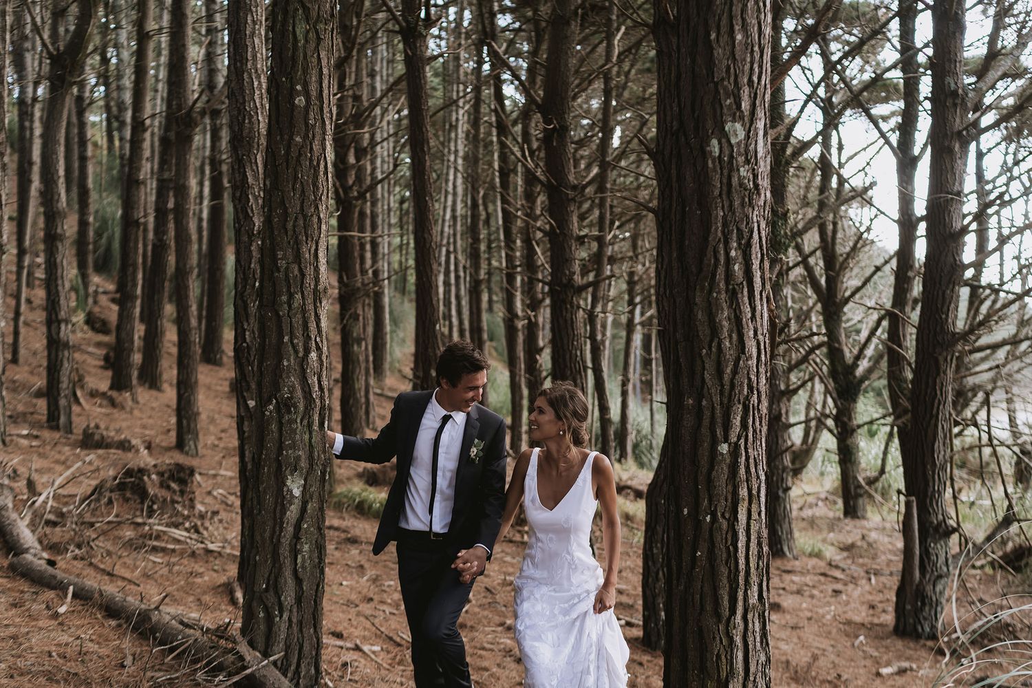 A couple in formal attire walks among tall pine trees during what appears to be an outdoor wedding photo shoot.