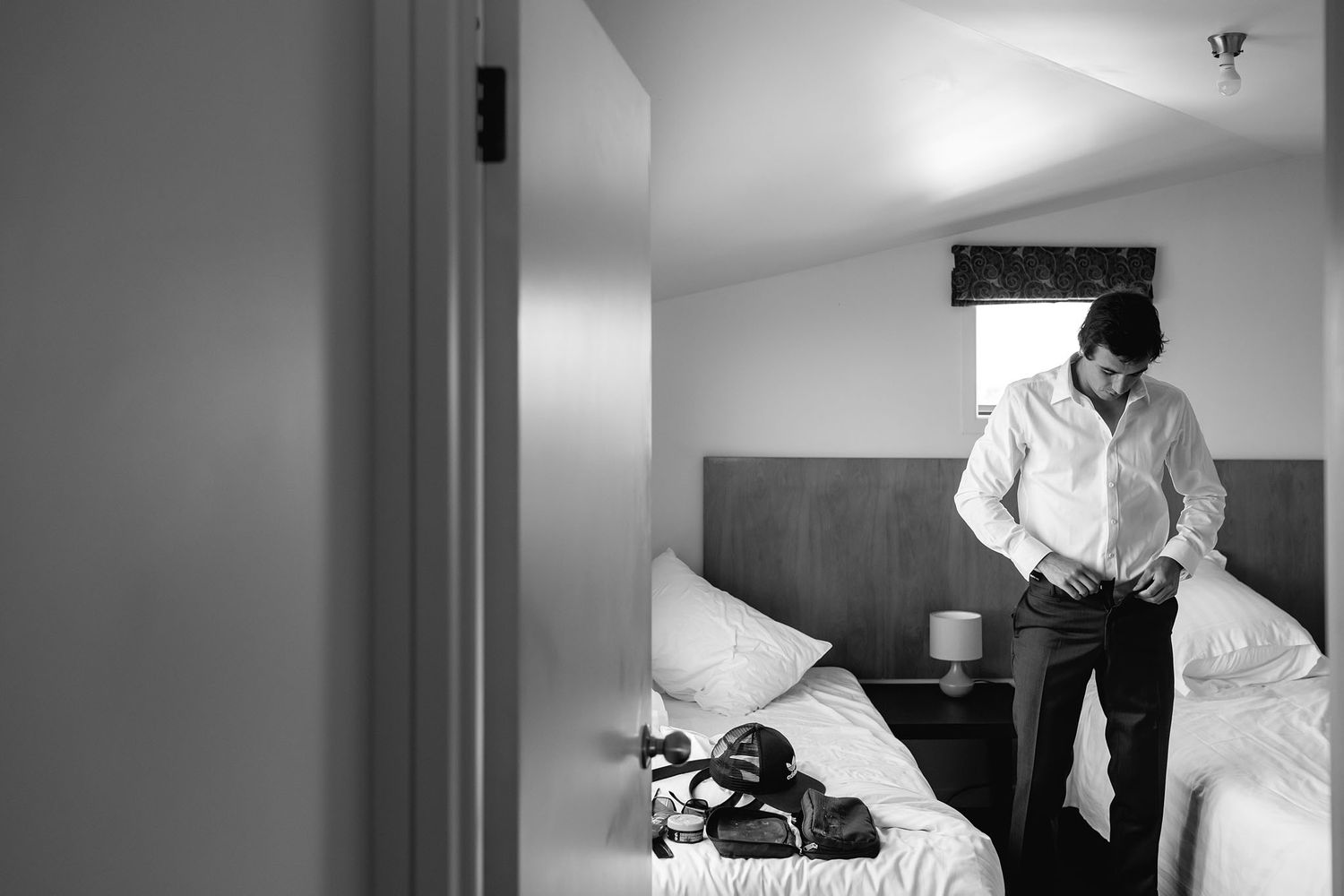 A person wearing a white shirt stands in a minimalist hotel room with a bed.