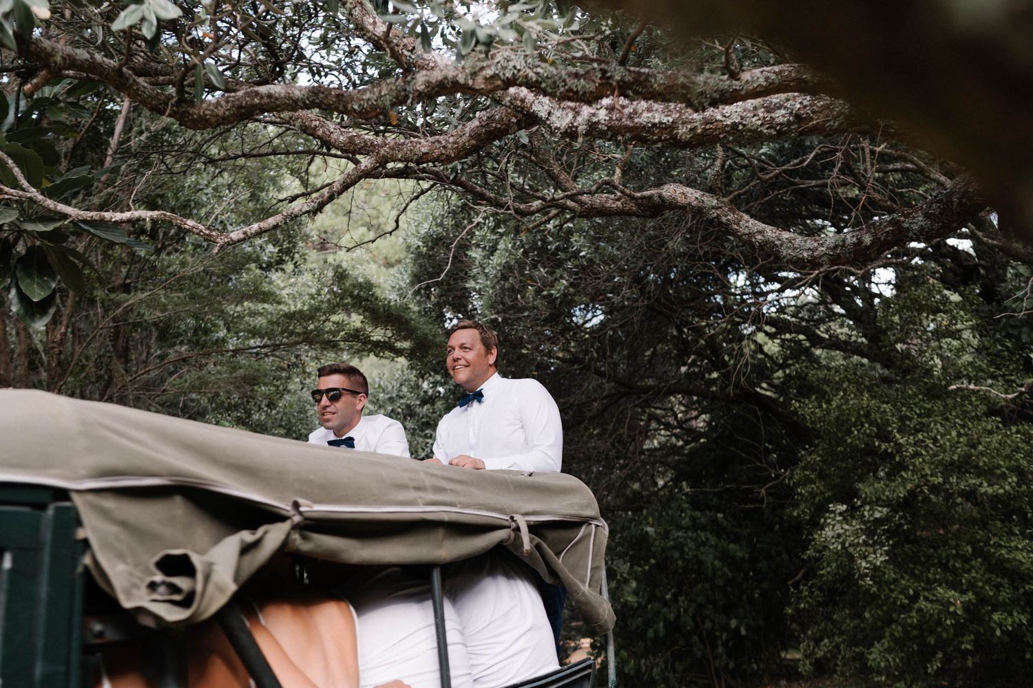 Two people stand in a covered golf cart beneath tree branches on a wooded path.