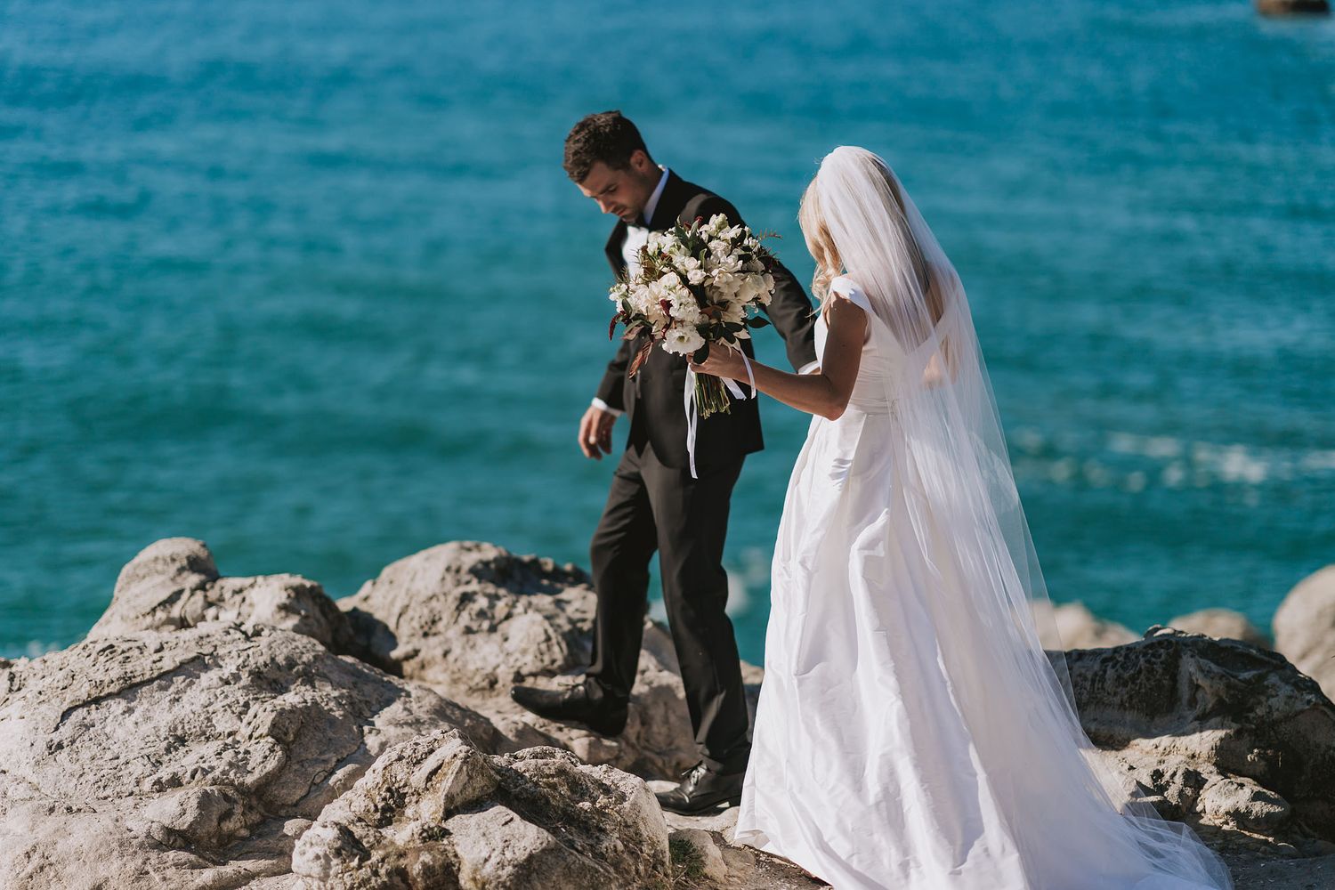 Newlywed couple stands together on coastal rocks against bright turquoise ocean waters during outdoor wedding ceremony.