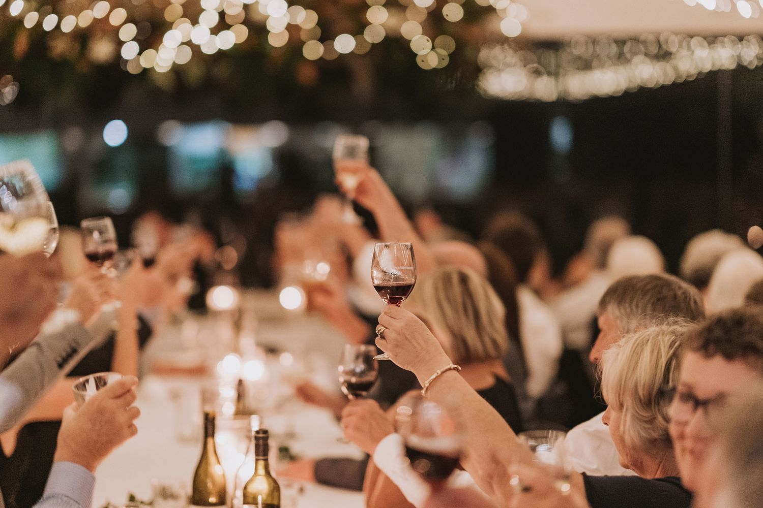 People raise wine glasses in celebration at an outdoor evening gathering with string lights overhead.