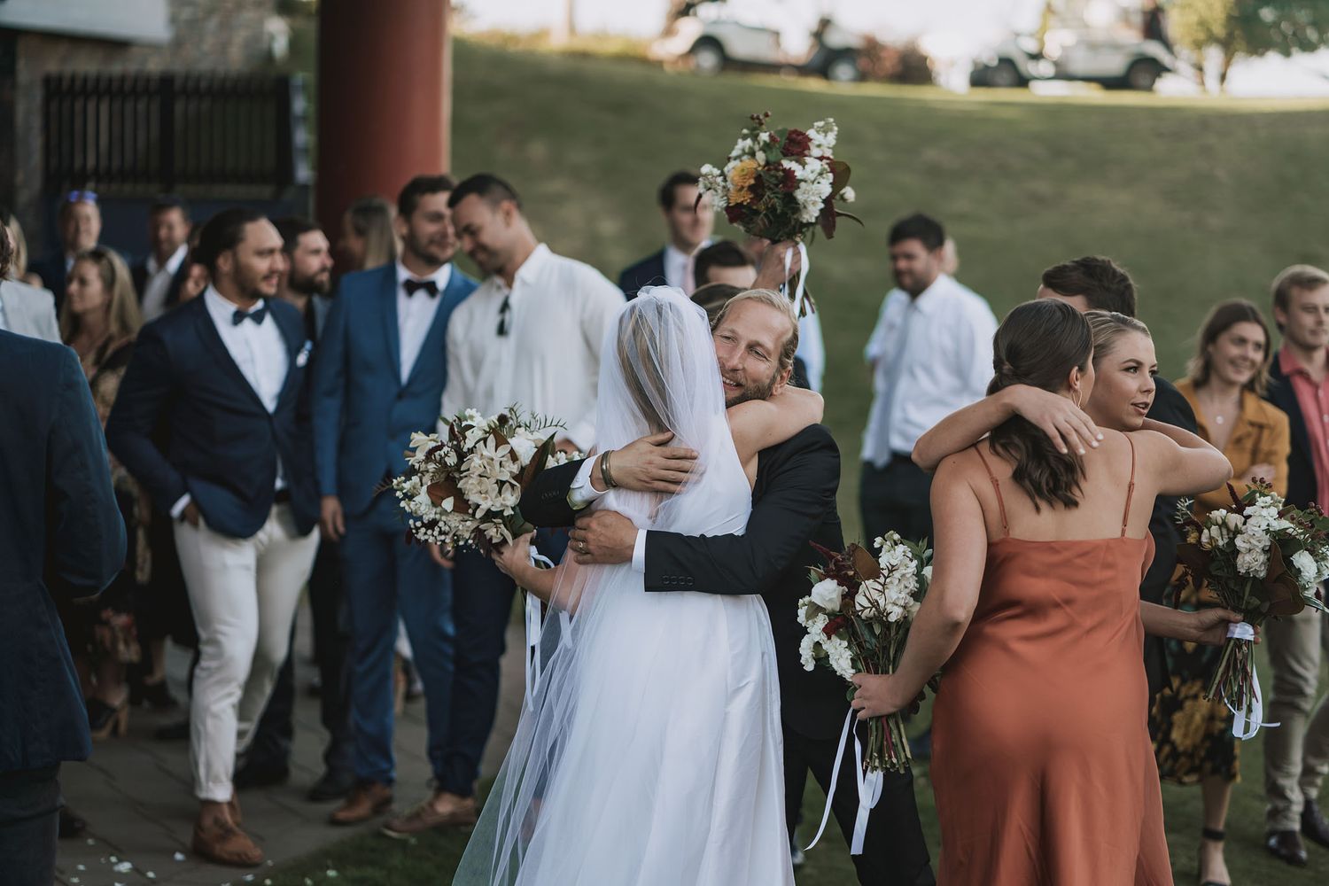 Wedding party in formal attire gathers outdoors with bridesmaids holding floral bouquets during sunset celebration.