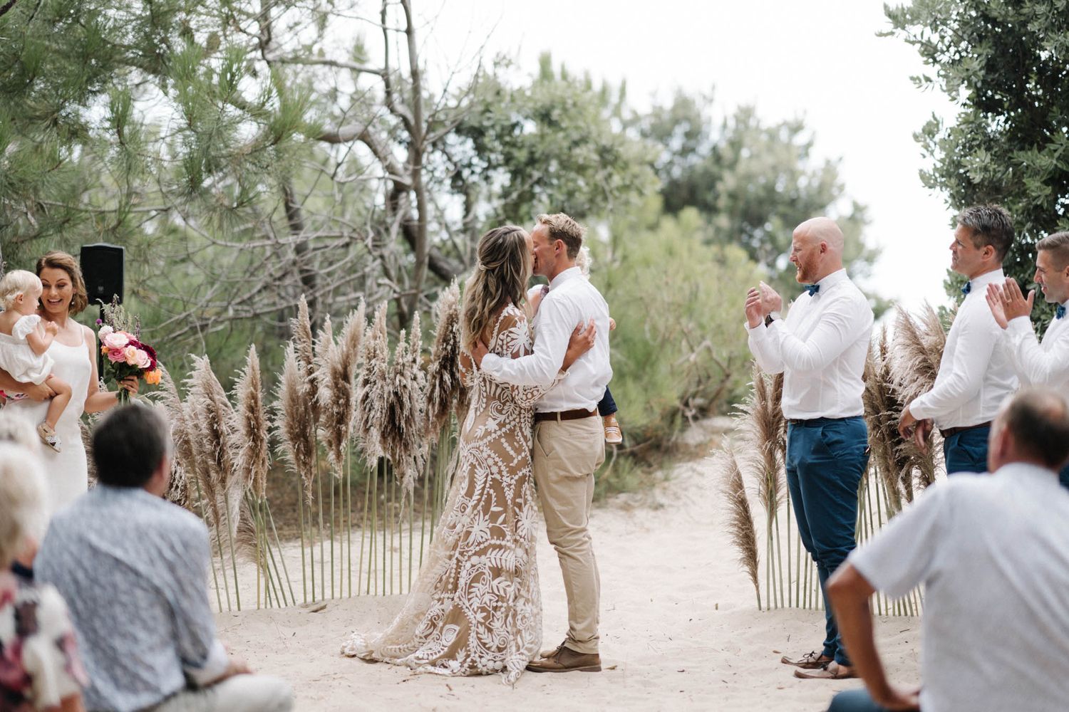 Intimate outdoor wedding ceremony with dried pampas grass decorations on a sand path among olive trees.