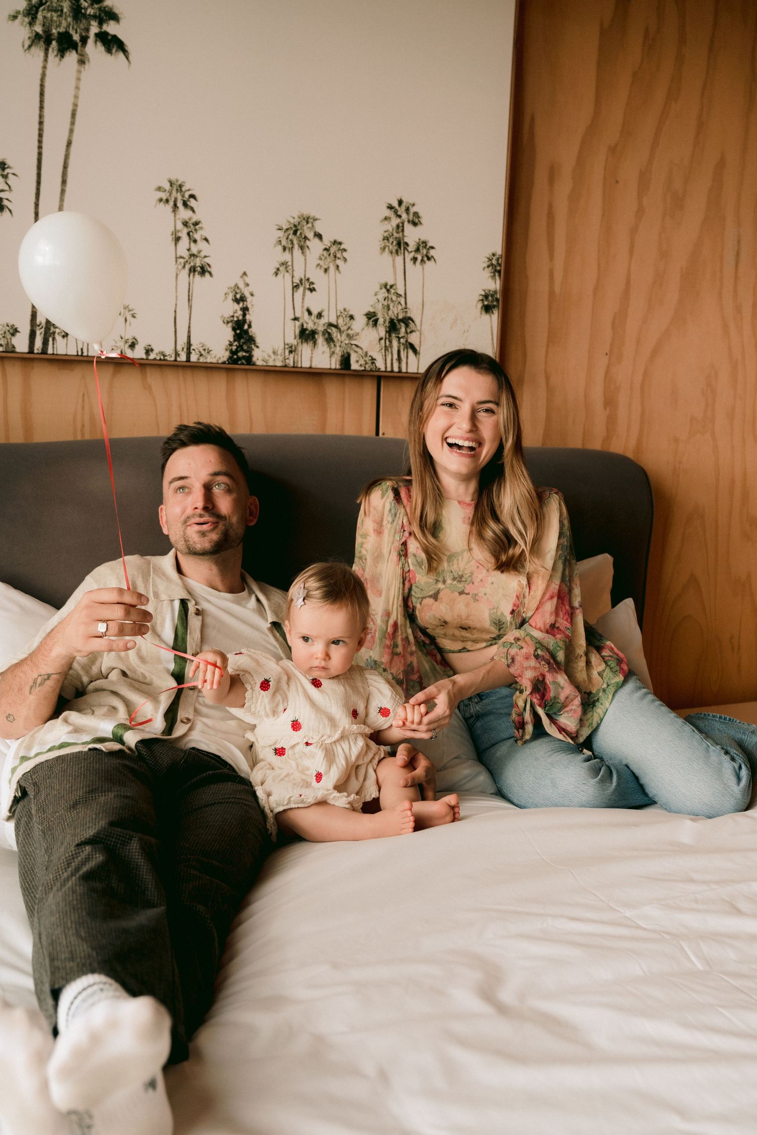 A family relaxes together on a bed with matching floral outfits and a white balloon against a wooden wall with palm artwork.