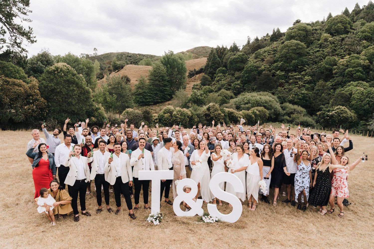 Large wedding party gathered outdoors on a grassy hillside for group photos.