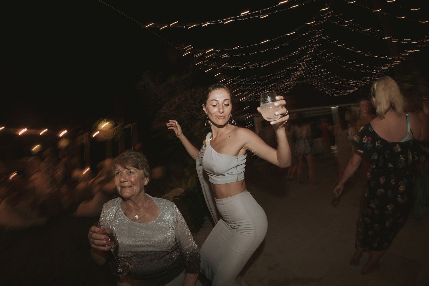 People dance and socialize at a dimly lit party venue with string lights creating a festive atmosphere.