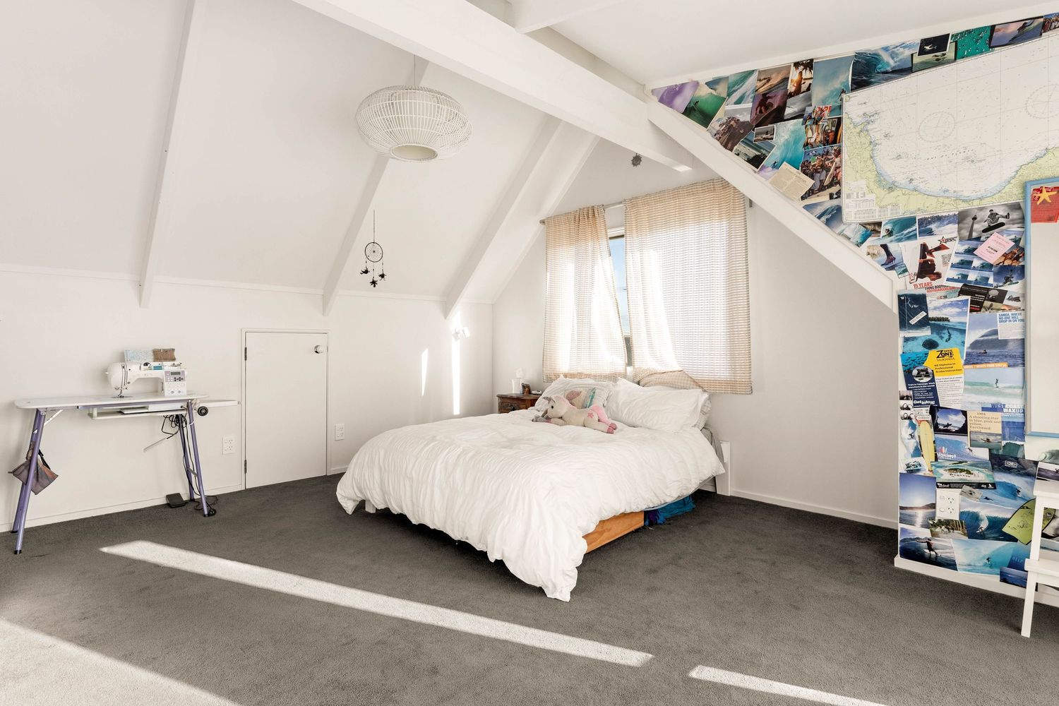 Modern attic bedroom with white bedding, exposed beams, and a photo collage wall features concrete flooring and natural light.