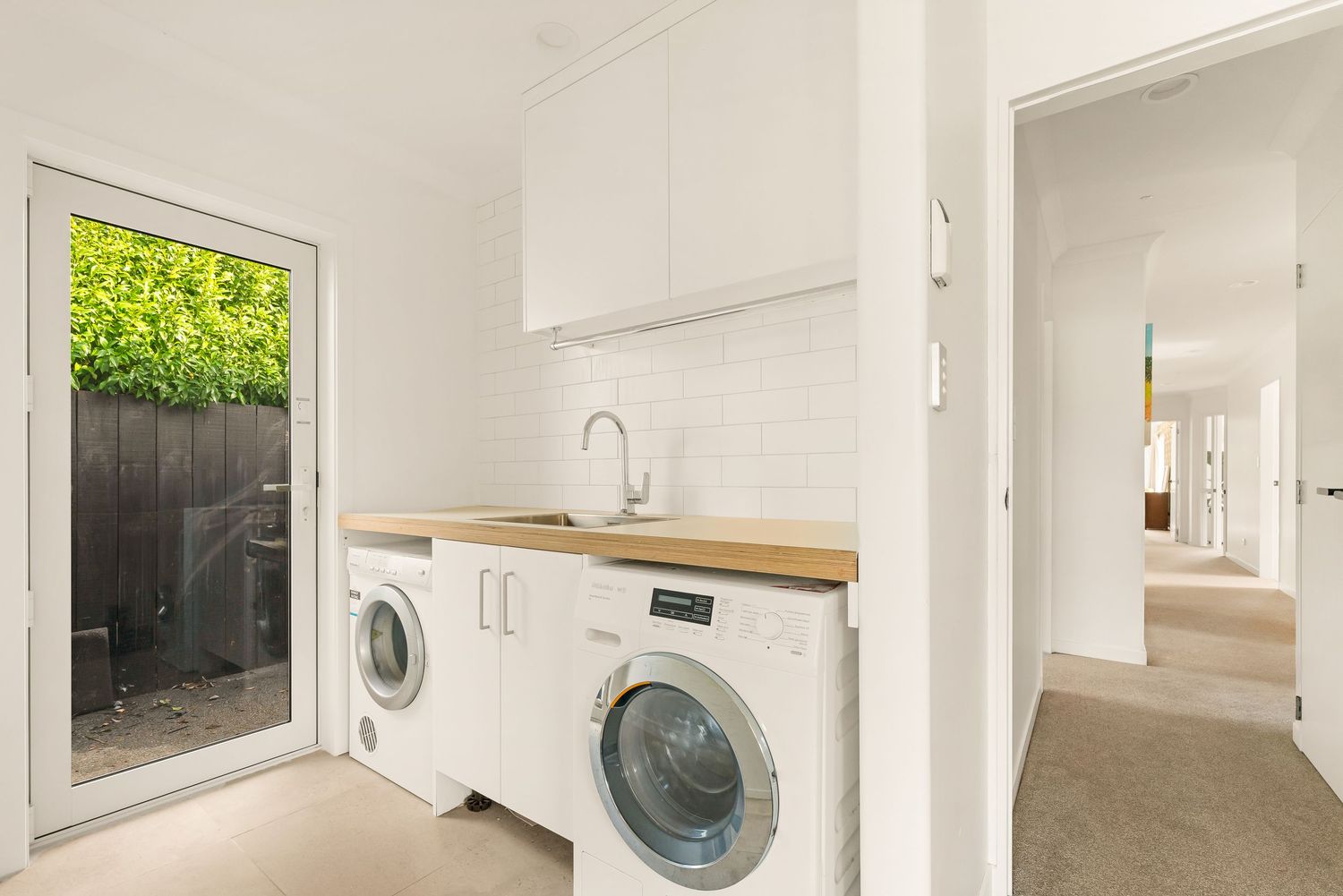 Modern laundry room with white cabinets, wooden countertop, and side door leading to outdoor garden.