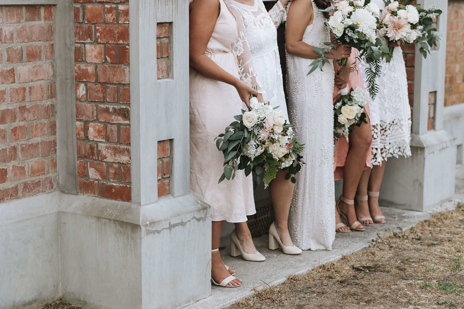 Bridesmaids in white dresses holding elegant bouquets of white flowers and greenery stand against a brick wall.