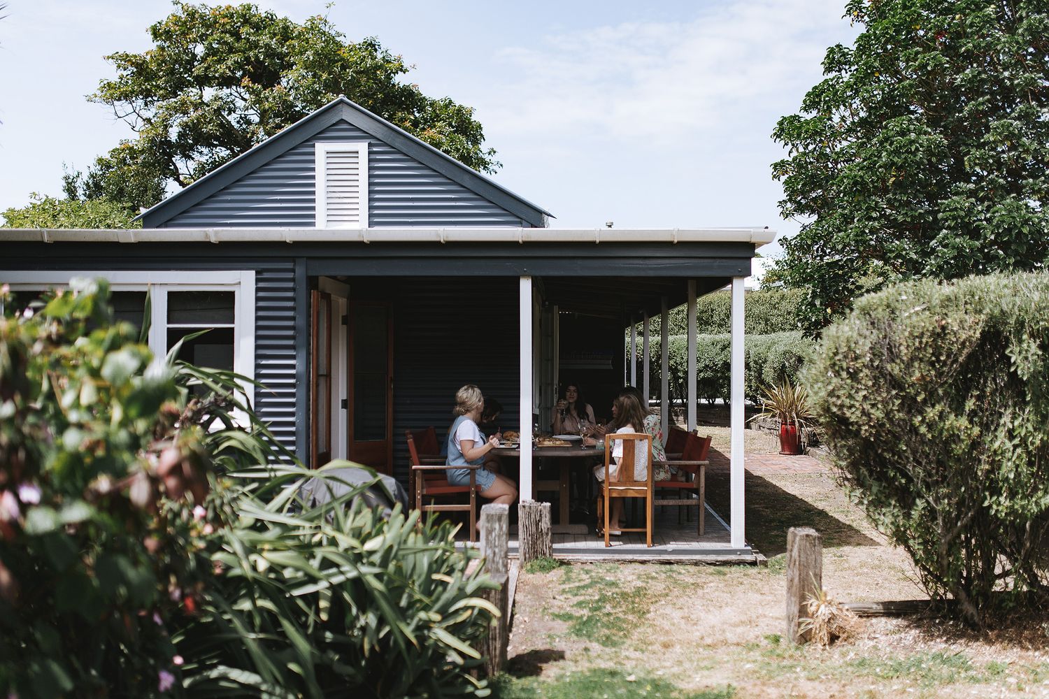 Blue-sided cottage with covered porch surrounded by lush garden landscaping on a sunny day.