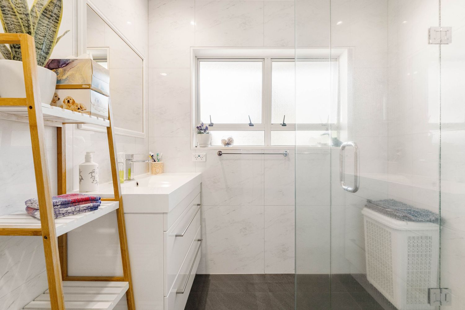 Clean white modern bathroom with glass shower enclosure, vanity storage, and bamboo ladder shelf for towels.