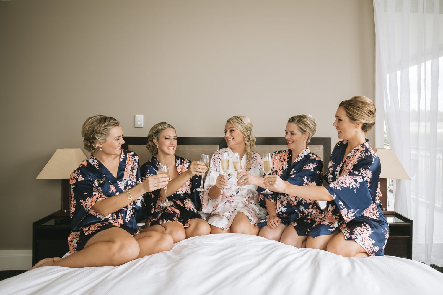 Wedding party sits in a row and poses for candid photographs showing laughter and joy during pre-ceremony preparations.