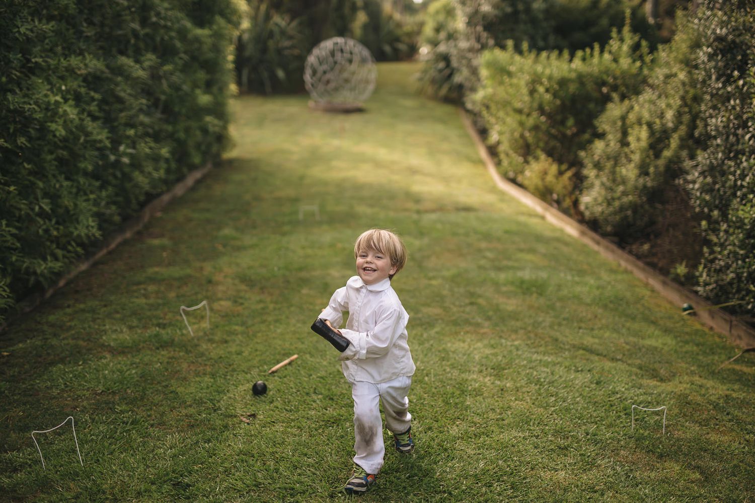A child in white clothing runs playfully through a sun-dappled garden path bordered by lush greenery.