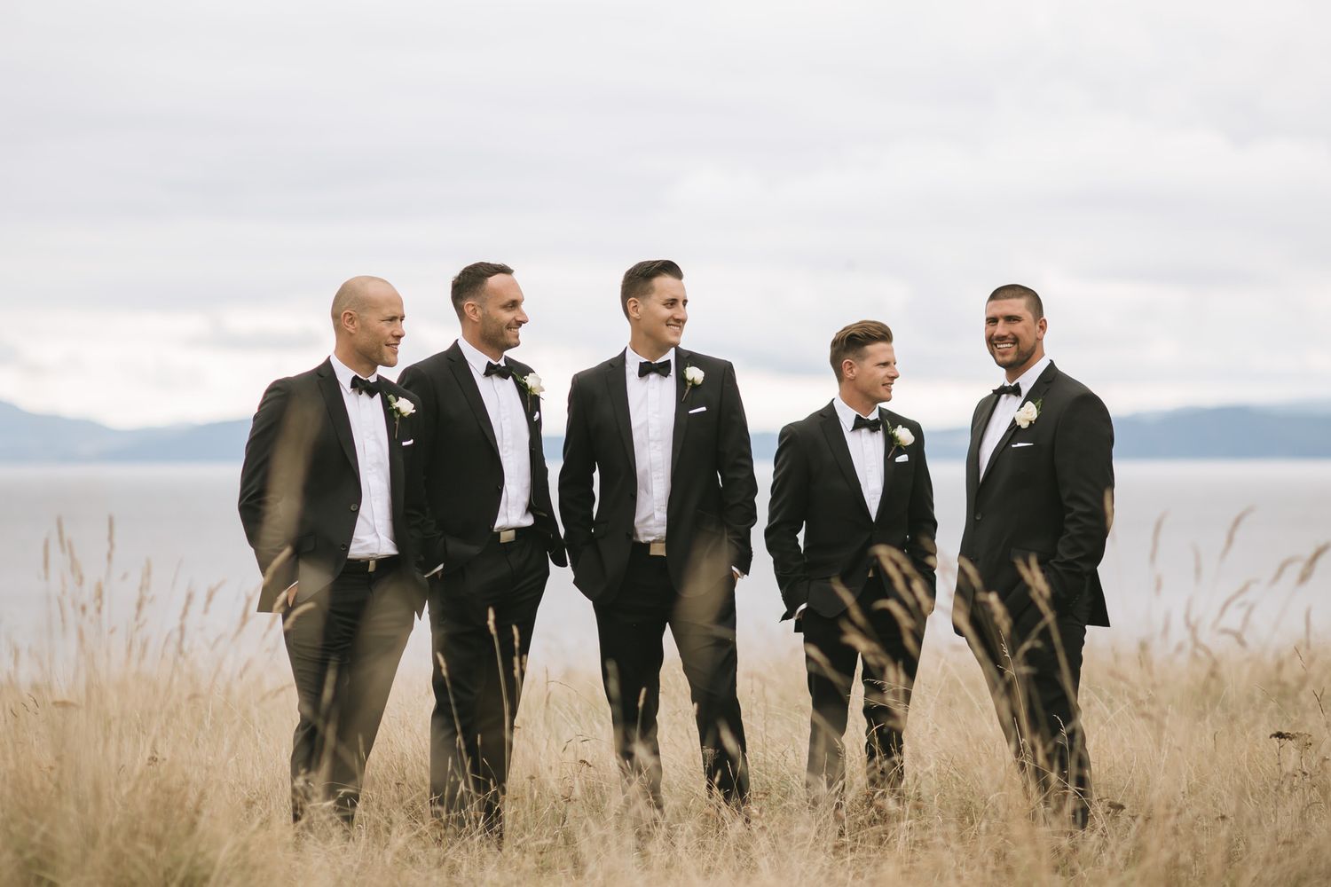 A group stands together in formal tuxedos against a scenic backdrop of tall grass and mountain views.