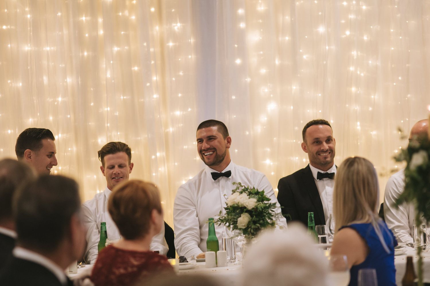 Wedding reception guests enjoying dinner and conversation at a table with twinkling fairy lights backdrop.