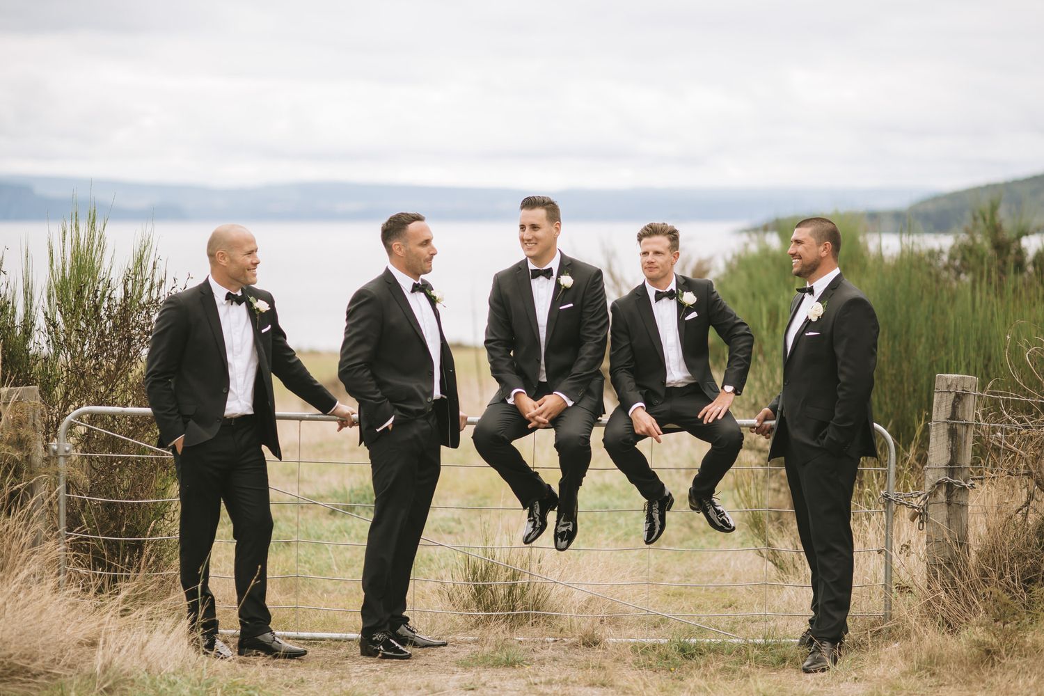 Groomsmen in black tuxedos jump playfully against a scenic outdoor backdrop, creating a fun and energetic wedding photo.