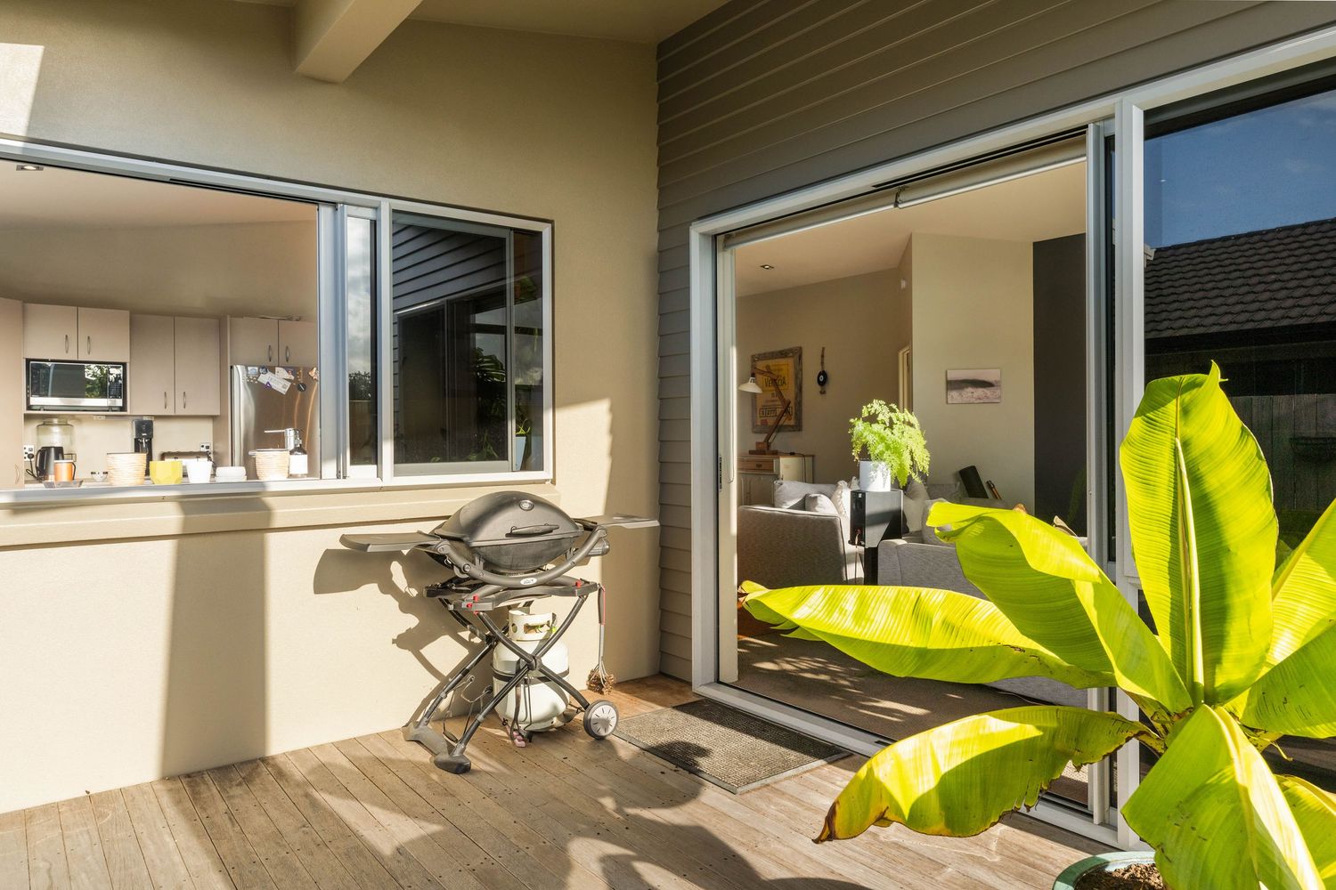 Modern home interior with sliding glass doors opening to a deck, bright green plant leaves in foreground and kitchen glimpse.