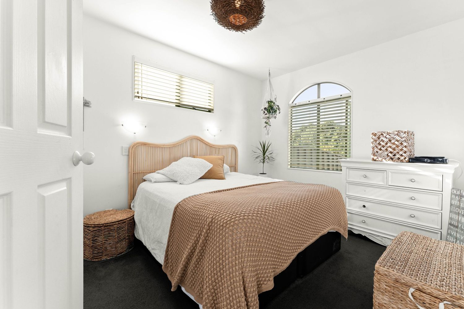 Light-filled bedroom featuring rattan furniture, beige bedding, and arched window with white dresser.