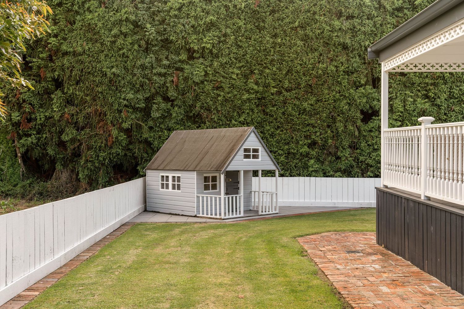 Modern backyard patio space with brick pavers and white fencing surrounded by greenery.