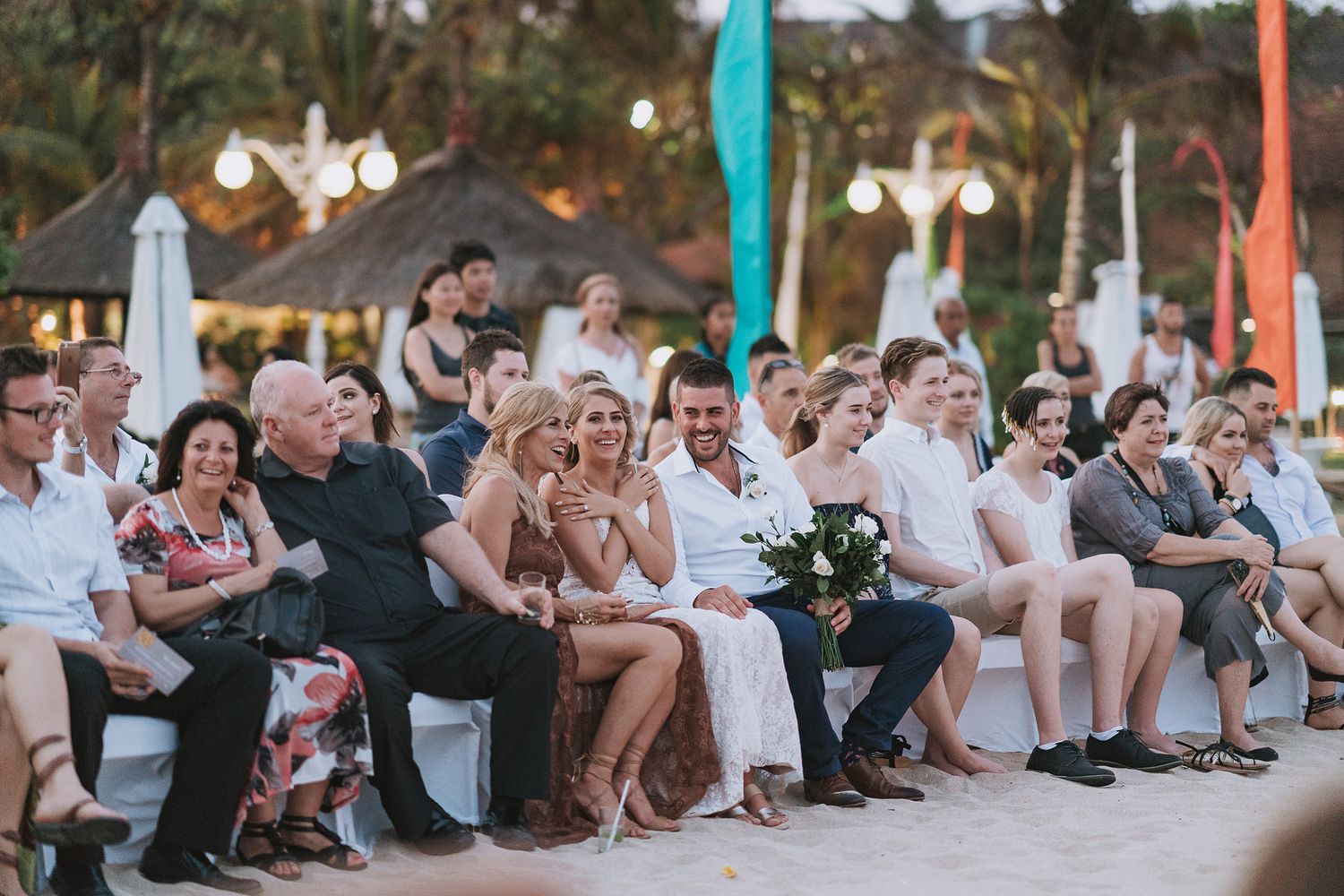 Wedding guests seated in white chairs watch an outdoor ceremony under strings of lights.