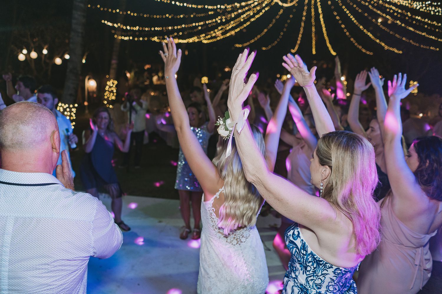 Wedding guests dance with raised arms under glowing string lights at an outdoor reception.