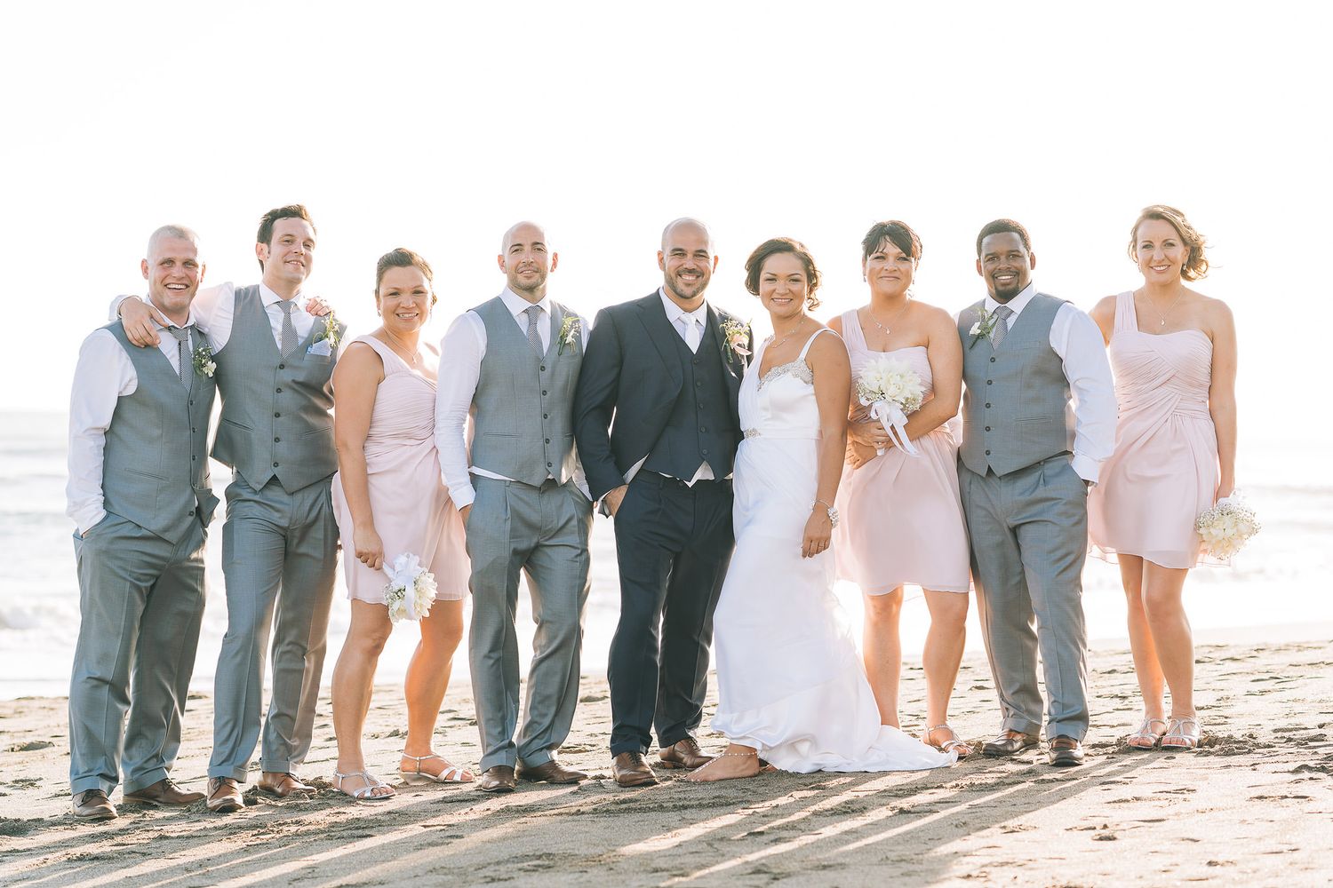 Wedding party posing together on a sunny beach in coordinated pink and gray outfits.