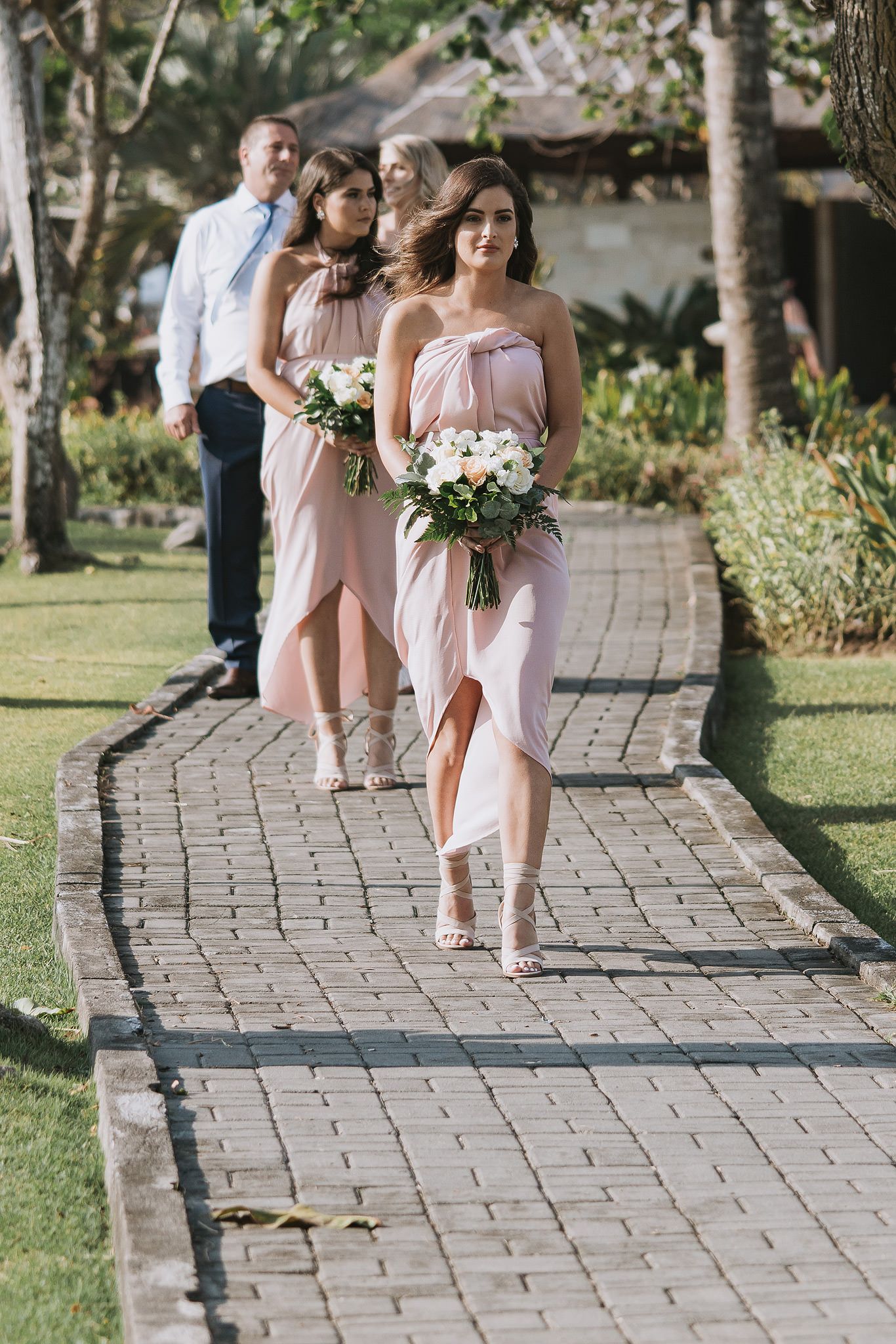 Bridesmaids in blush pink dresses walk down a stone path carrying white bouquets at an outdoor ceremony.