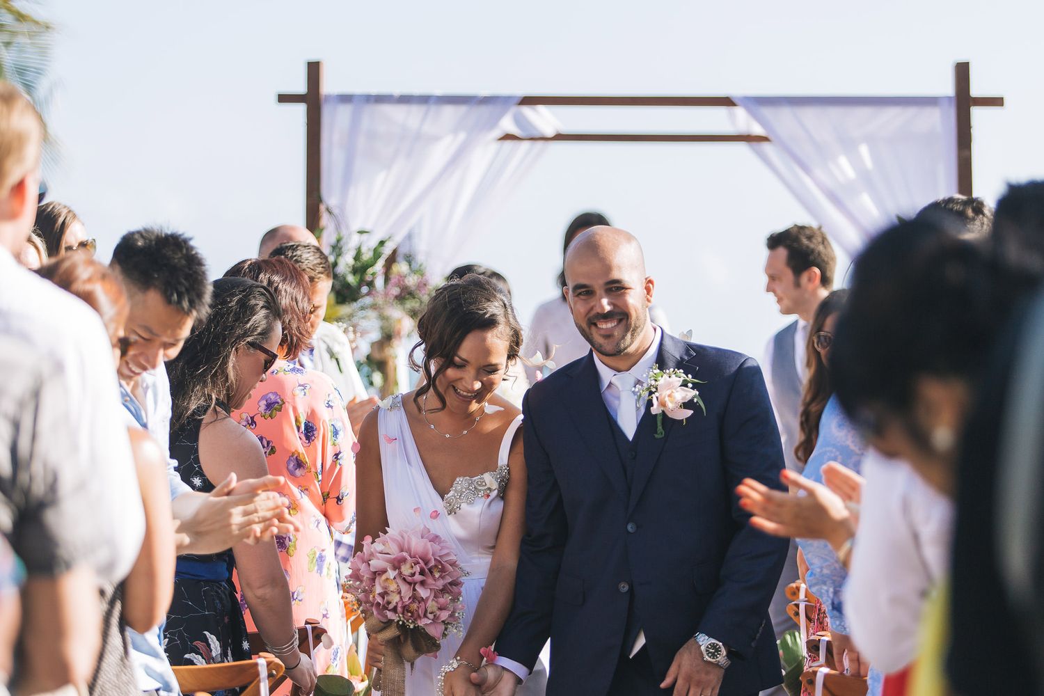 Guests celebrate and throw confetti at an outdoor wedding ceremony under blue skies.