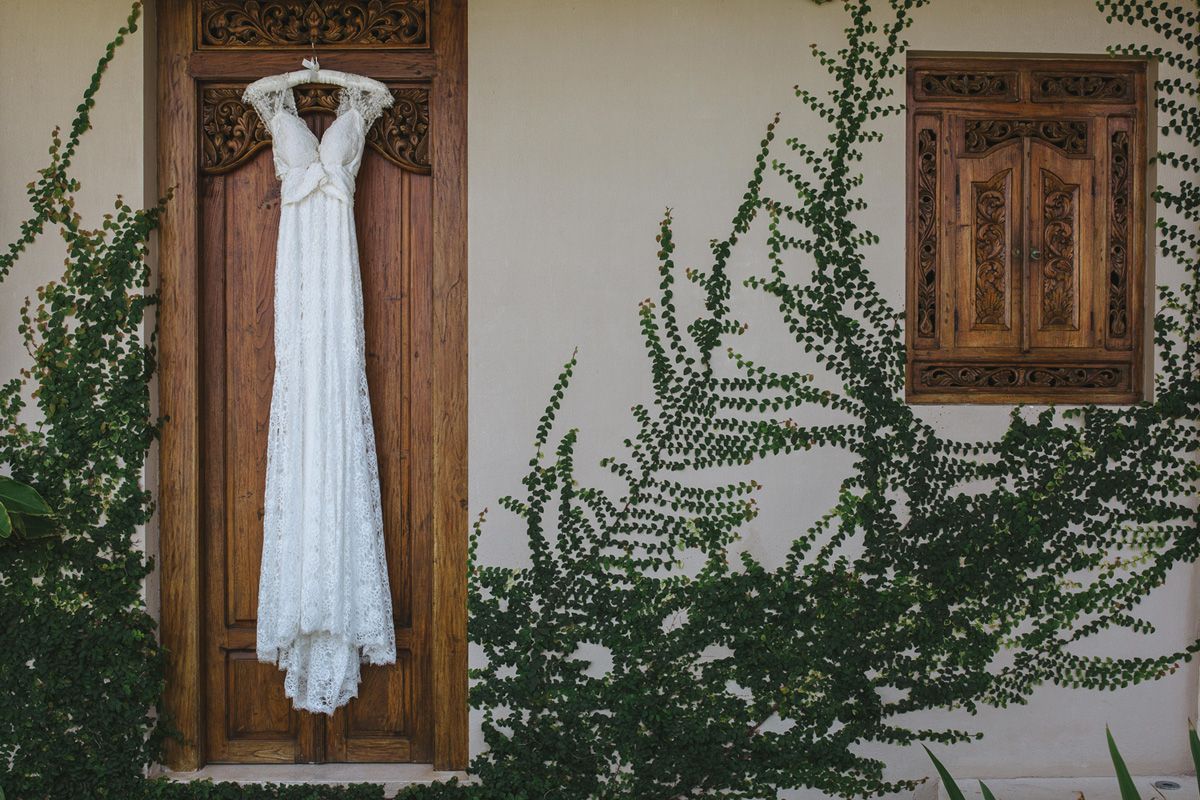 A white lace wedding dress hangs on a wooden door next to green climbing vines against a white wall.