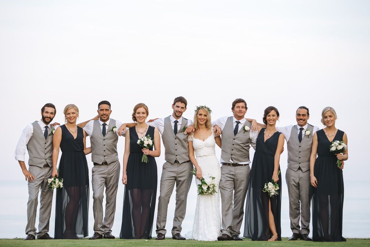 Wedding party in gray suits and navy dresses poses together on hilltop against sky background.