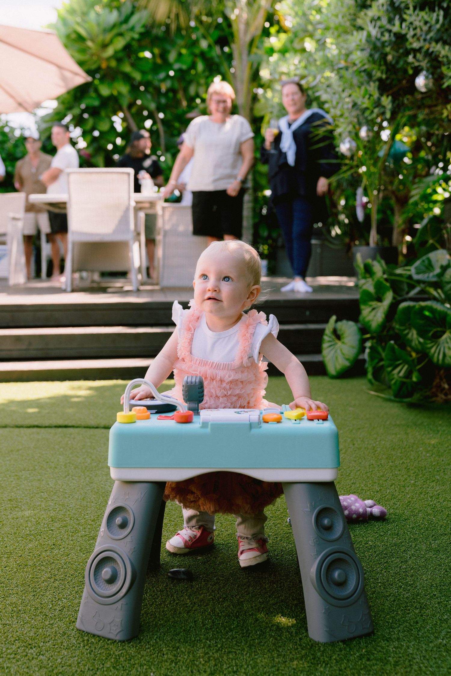 A baby in pink dress sits in a teal-colored play table on green turf with people gathering in the background.