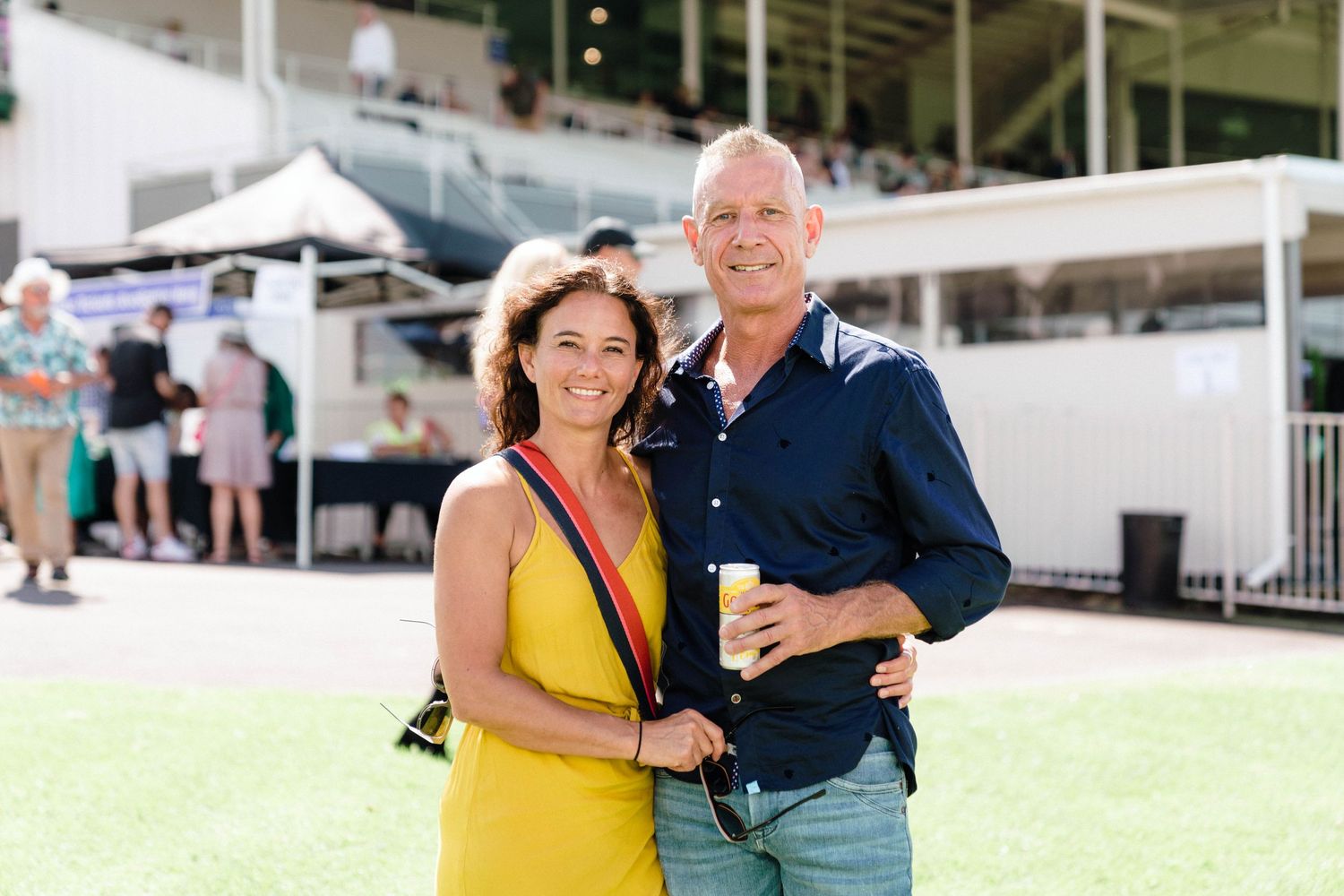 Two people dressed in summer attire pose together on grassy area at outdoor event venue with tents in background.