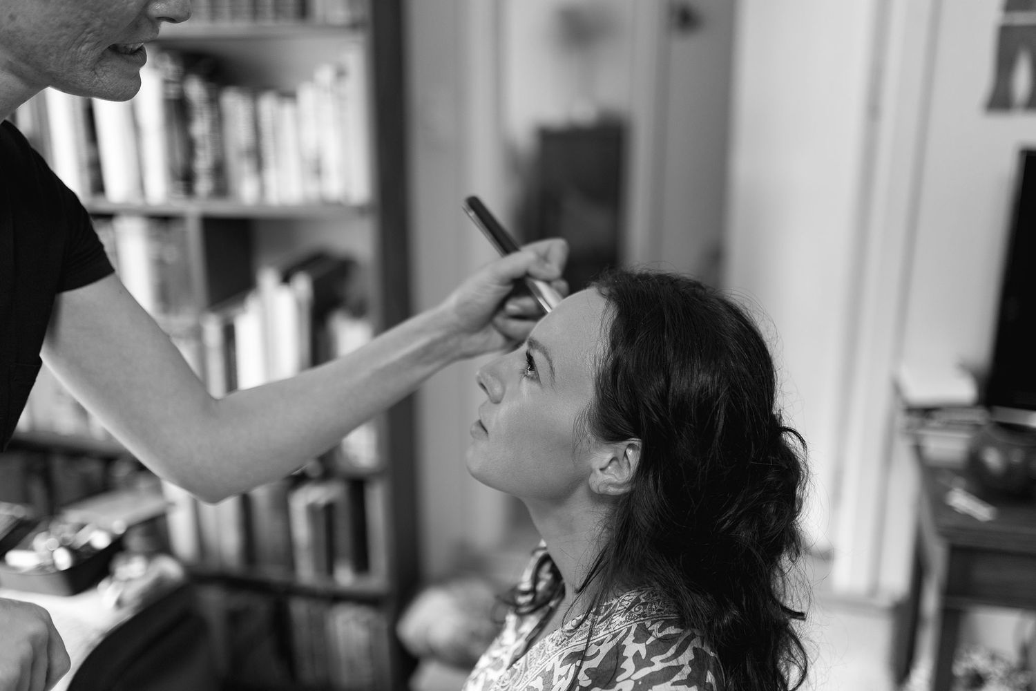 A black and white photograph showing makeup application being done in an indoor setting.