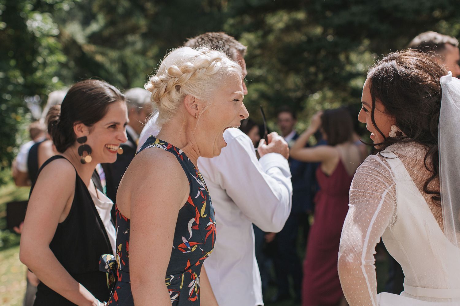 A group of guests share a joyful moment of laughter outdoors during a summer wedding celebration.