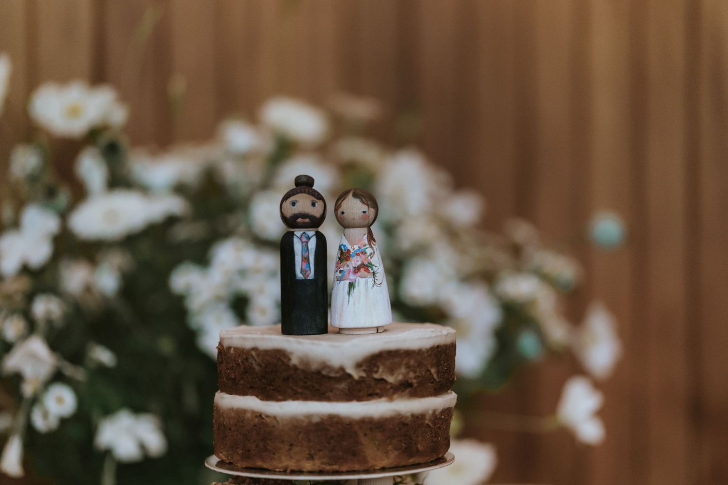 A rustic naked wedding cake decorated with cute bride and groom cake toppers surrounded by white flowers.
