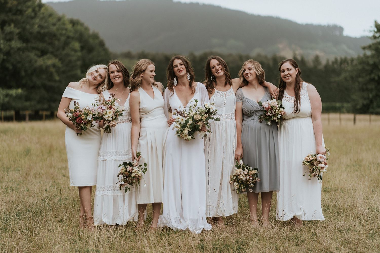 A wedding party wearing flowing white dresses stands together in a mountain meadow while holding floral bouquets.